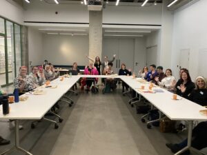 Image: A meeting room with two long tables arranged in a U-shape. Around the tables sit approximately 15-20 people, many of whom are waving at the camera.